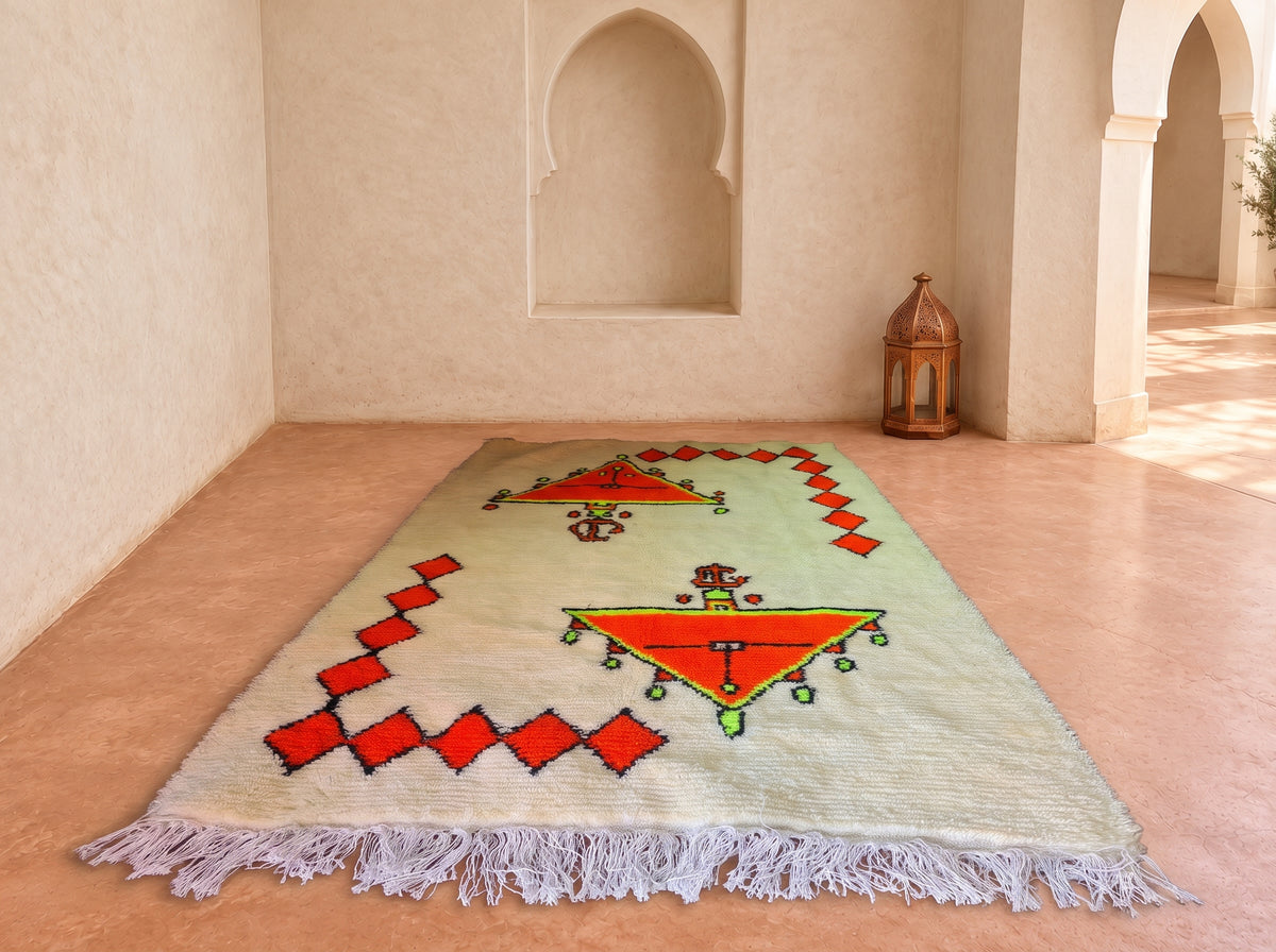 A rectangular Moroccan-style wool rug with a light sage green base, featuring a central red and orange geometric motif, red diamond borders, and white fringe, displayed on a terracotta floor in a Moroccan-style room with arches.