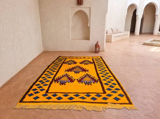 A vibrant yellow Moroccan-style rug with geometric patterns and fringe, displayed on a terracotta floor in a minimalist courtyard with white walls and arches.