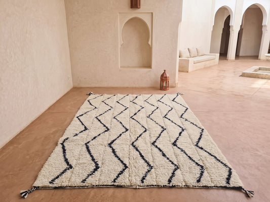A large rectangular Moroccan-style rug with a black zigzag pattern on a cream bouclé texture, displayed on a terracotta floor in a minimalist, sunlit courtyard with arches and a bench in the background.