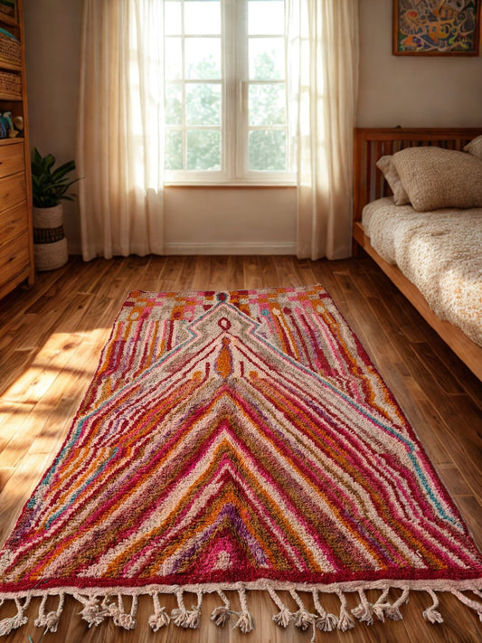 Colorful geometric-patterned rug on a wooden floor with a window and bed in the background