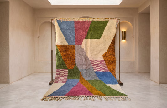 A large multicolor geometric area rug displayed on a black metal stand in a minimalist beige room with arched doorways and a skylight.